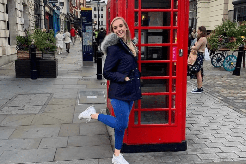 A woman in a black jacket stands beside a red telephone booth, smiling in an urban setting.