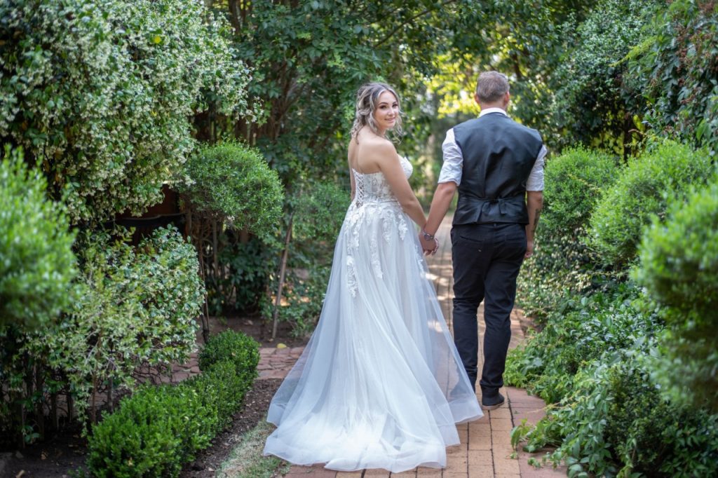 A bride and groom stroll hand in hand along a garden path, surrounded by lush greenery.