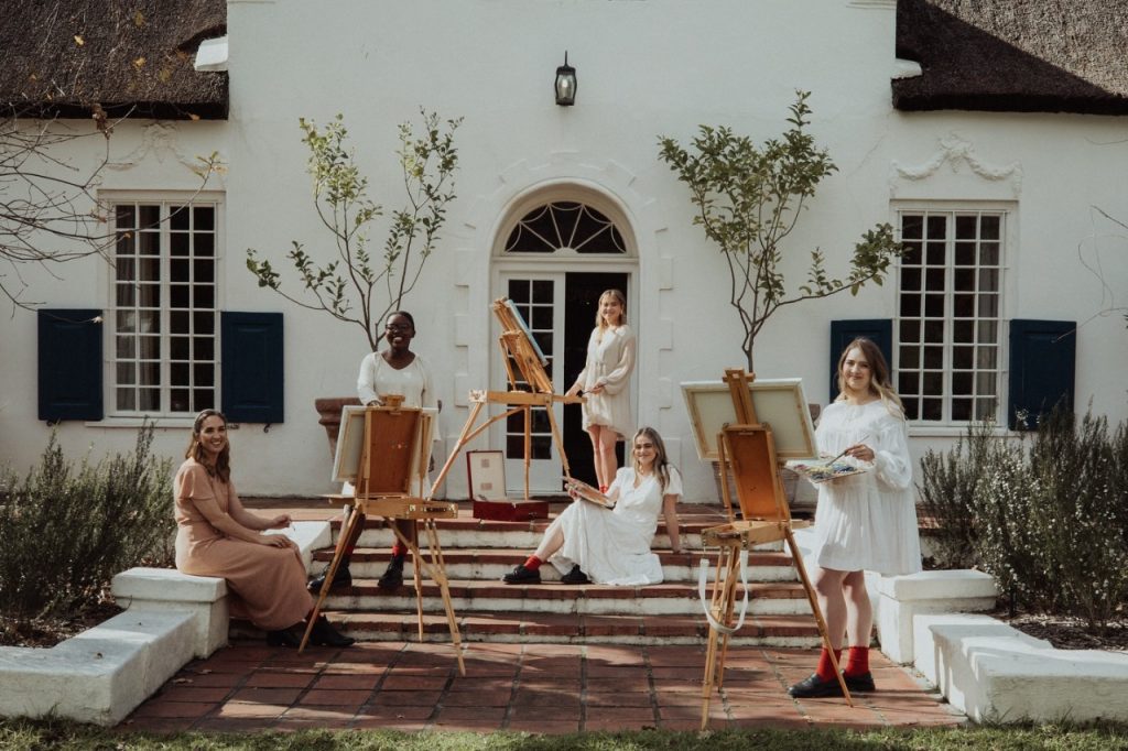 Five women painting on easels in front of a house.