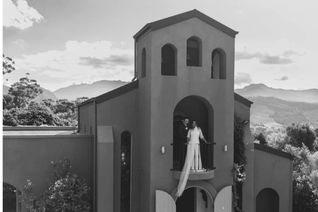 Bride and groom standing together on a balcony, smiling and embracing, with a scenic view in the background.