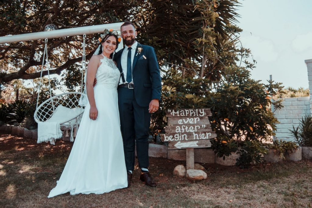 A bride and groom smile together for a photo, standing in front of a decorative swing in a picturesque outdoor setting.